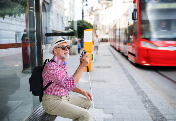 A senior blind man with white cane waiting for public transport in city.