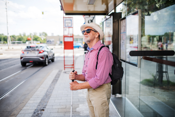 A senior blind man with white cane waiting at bus stop in city.