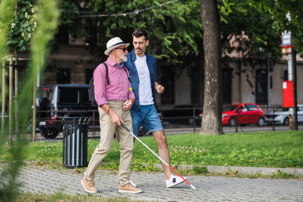 A young man and blind senior with white cane walking on pavement in city, talking.