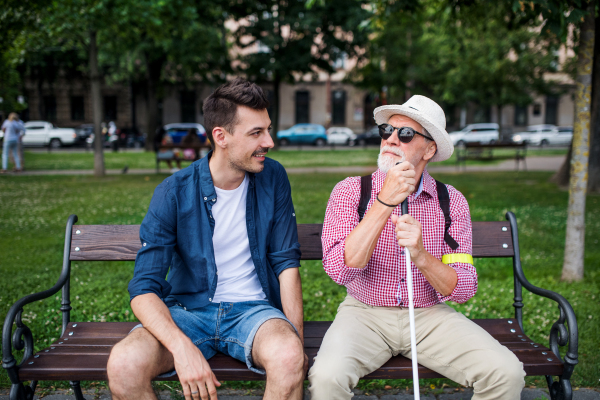 A young man and blind senior with white cane sitting on bench in park in city, talking.