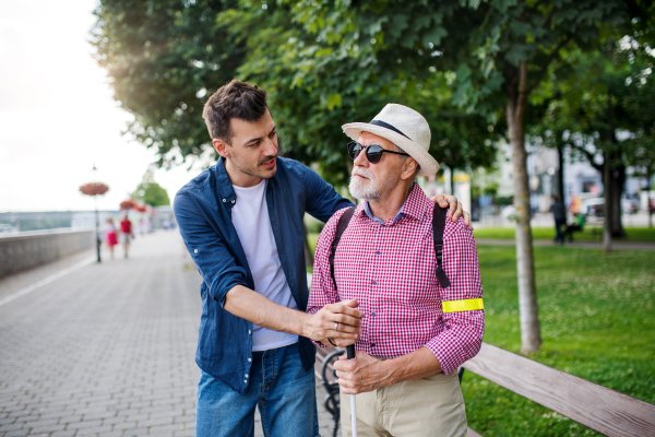 A young man and blind senior with white cane walking on pavement in city, talking.