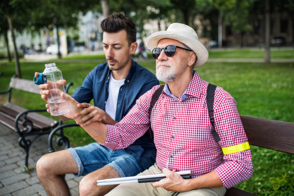 A young man and blind senior with white cane and water bottle sitting on bench in park in city.