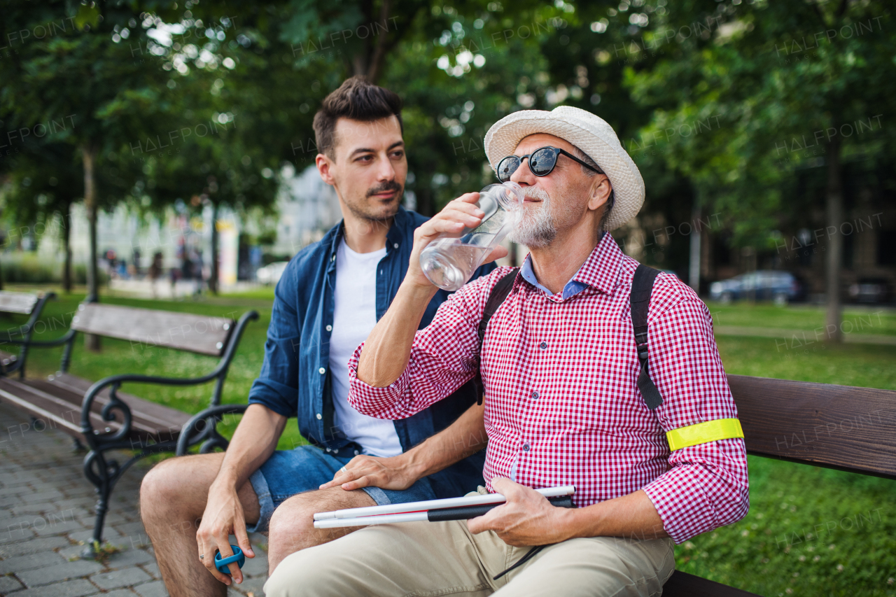 A young man and blind senior with white cane sitting on bench in park in city, drinking water.