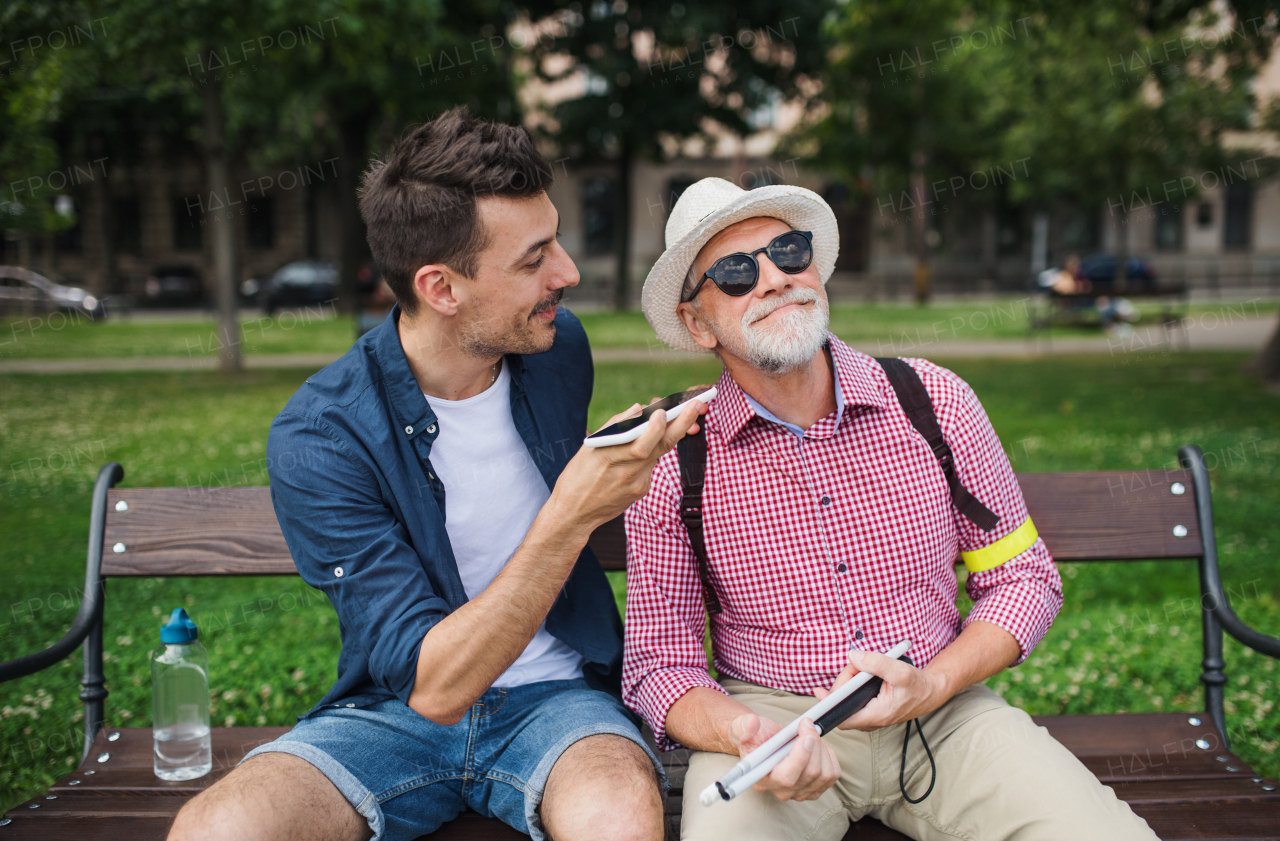 A young man and blind senior sitting on bench in park in city, using smartphone.