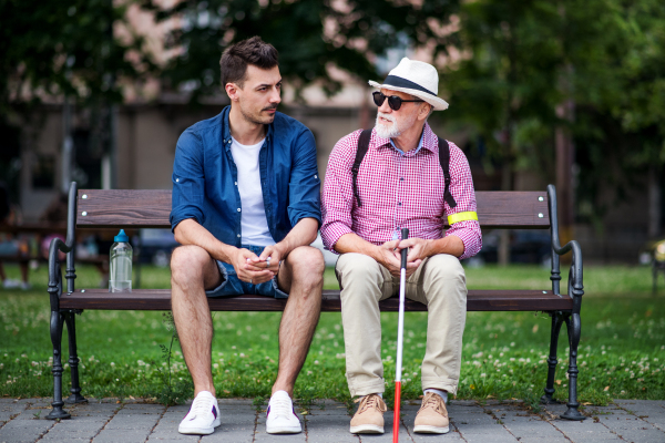 A young man and blind senior with white cane sitting on bench in park in city.