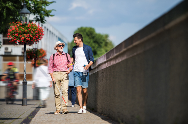 A young man and blind senior with white cane walking on pavement in city, talking.