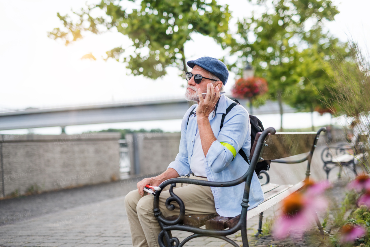 A senior blind man with smartphone sitting on bench in park in city.