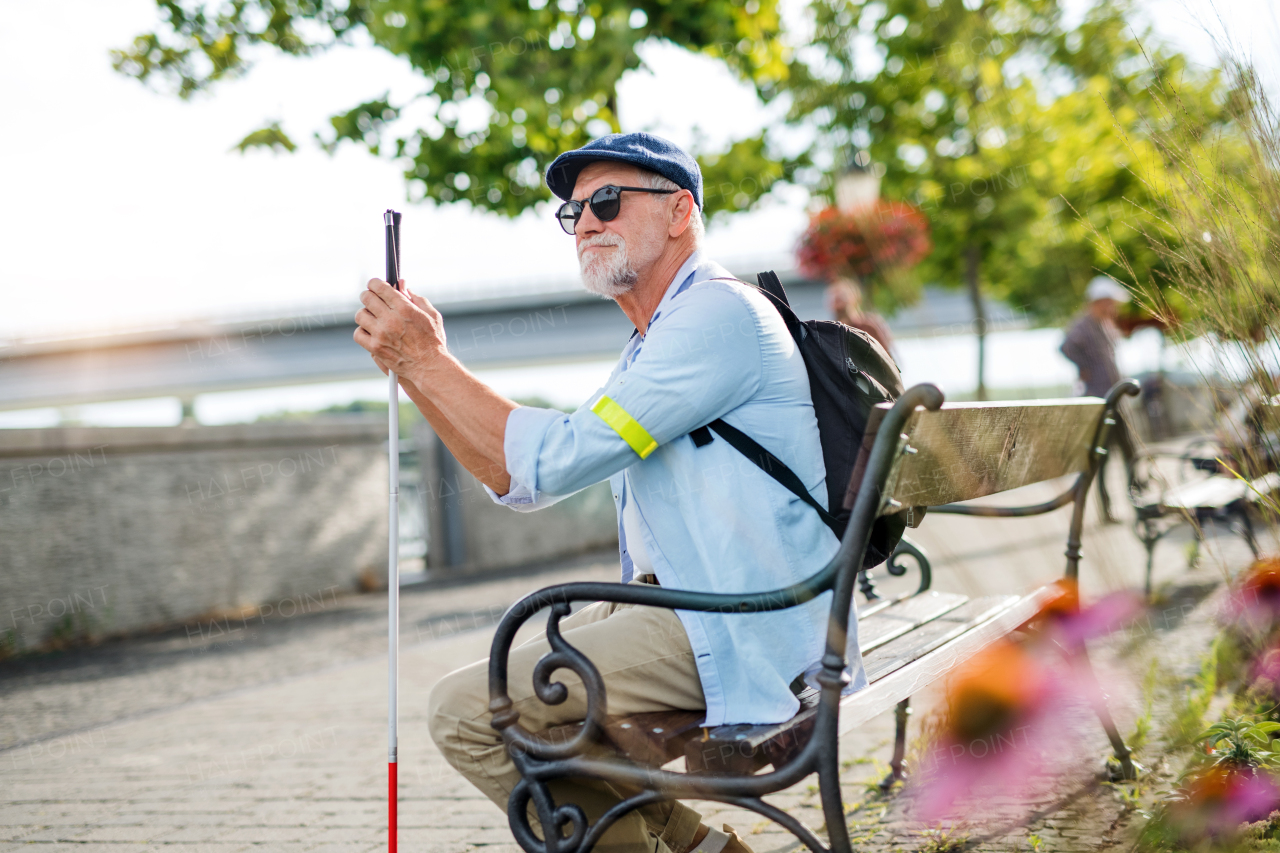 A senior blind man with white cane sitting on bench in park in city.