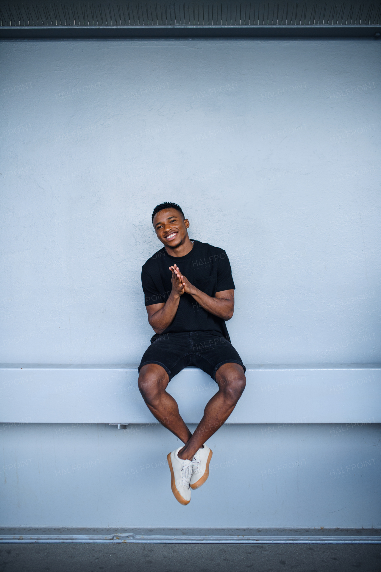 Cheerful young black man sitting against wall background, looking at camera.