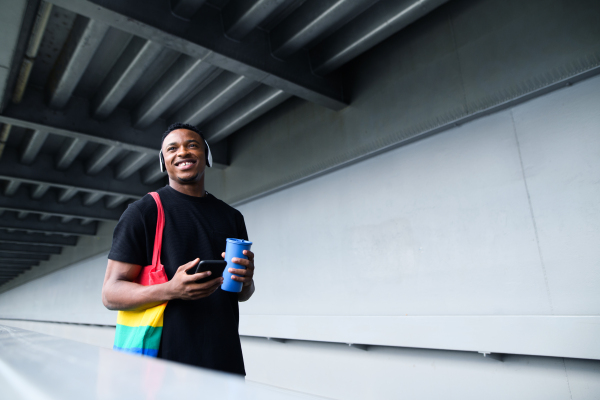 Cheerful young black man commuter outdoors in city, walking with headphones and smartphone.