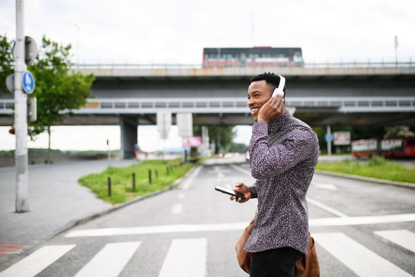 Cheerful young black man commuter outdoors in city, walking with headphones and smartphone.