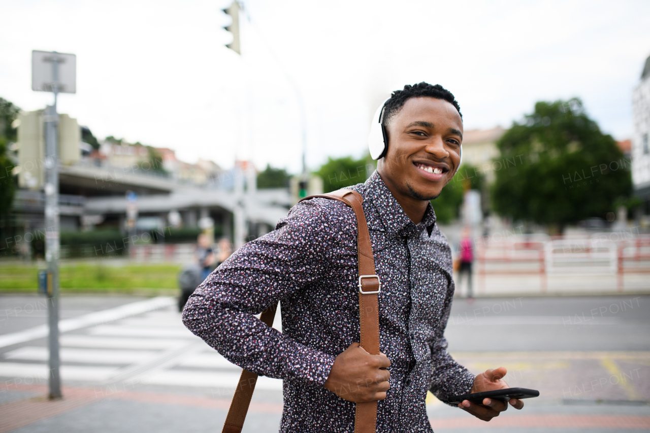 Cheerful young black man commuter outdoors in city, walking with headphones and smartphone.