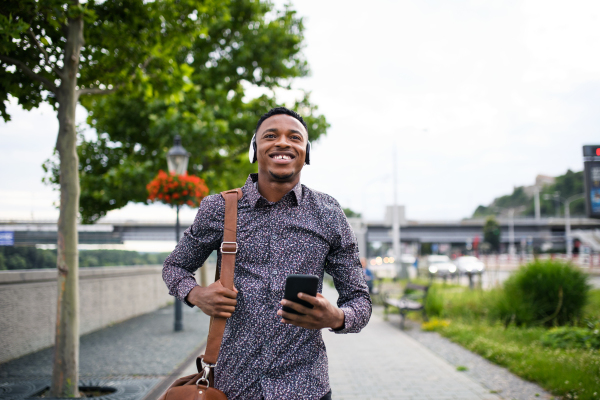 Cheerful young black man commuter outdoors in city, walking with headphones and smartphone.