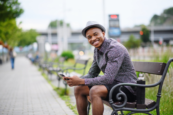 Cheerful young black man commuter sitting on bench outdoors in city, laughing.