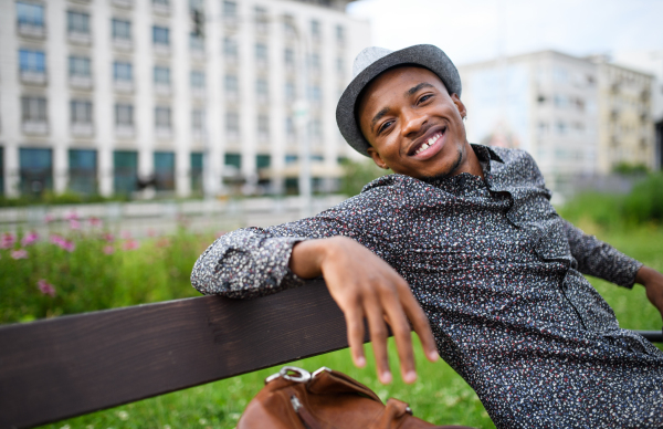 Cheerful young black man commuter sitting on bench outdoors in city, resting.