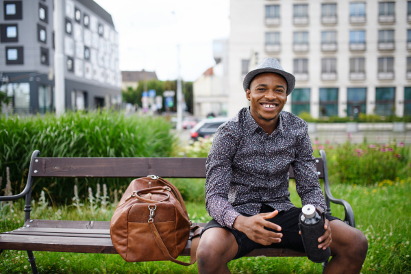 Cheerful young black man commuter sitting on bench outdoors in city, resting.