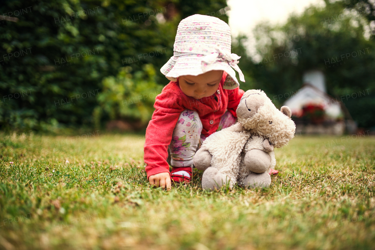 A front view of toddler girl standing outdoors on grass in garden in summer.