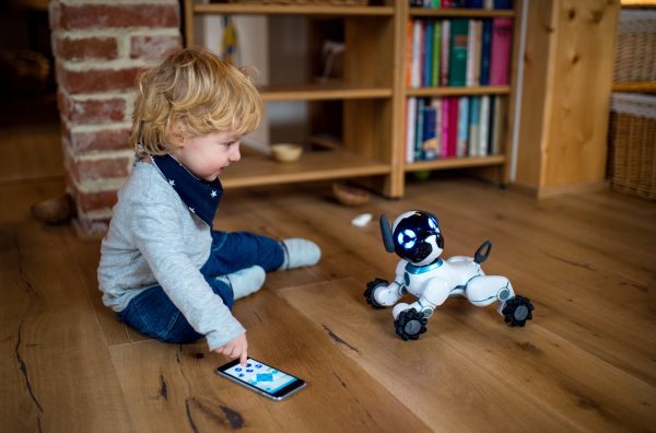 A happy cute toddler boy sitting indoors at home, playing with robotic dog.