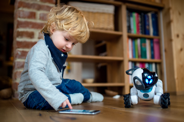 A happy cute toddler boy sitting indoors at home, playing with robotic dog.