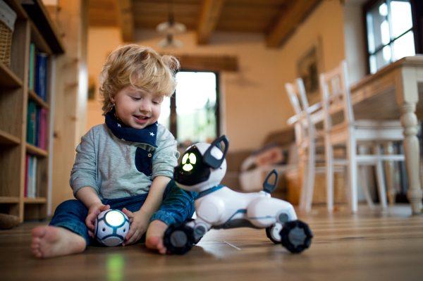 A happy cute toddler boy sitting indoors at home, playing with robotic dog.