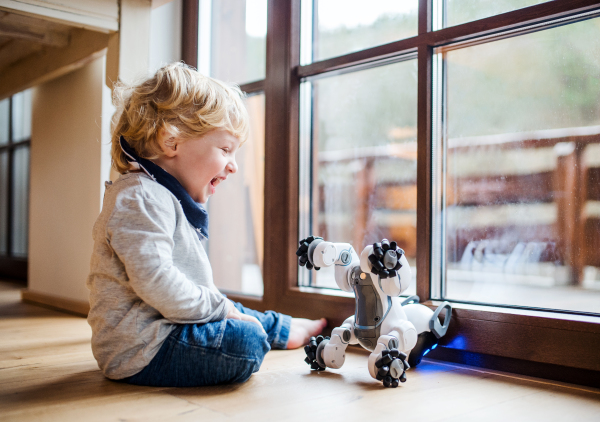 A happy cute toddler boy sitting on the floor indoors at home, playing with robotic dog.