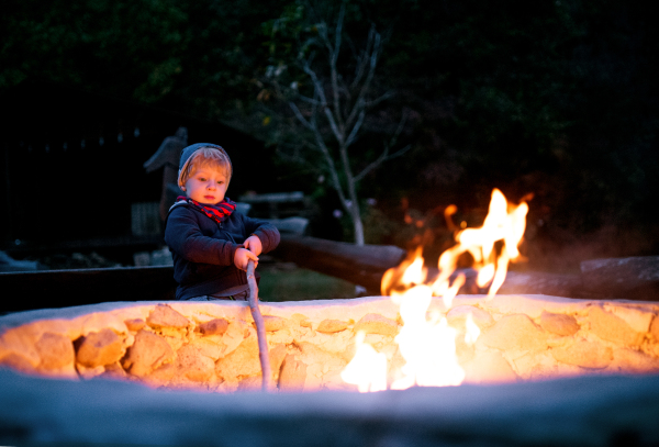 A toddler boy standing and playing by open fire in the evening outdoors in garden in summer.