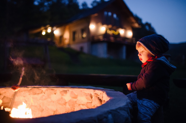 A toddler boy standing and playing by open fire in the evening outdoors in garden in summer.