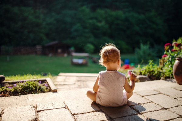 A rear view of toddler girl sitting outdoors on patio in summer, chalk drawing.