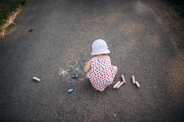 A rear view of cute toddler girl outdoors in countryside, chalk drawing on road. Copy space.