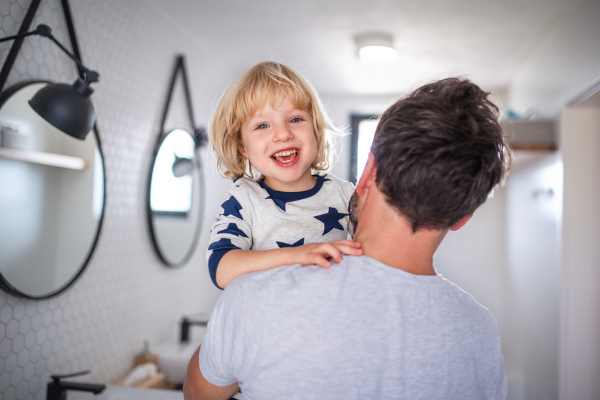 Mature father carrying small son indoors in bathroom, having fun.