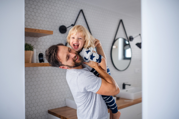 Mature father carrying small son indoors in bathroom, having fun.