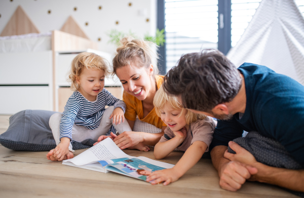 Front view of young family with two small children indoors in bedroom reading a book.