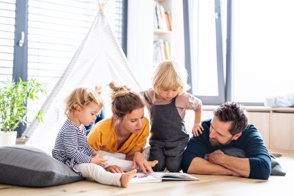 Front view of young family with two small children indoors in bedroom reading a book.