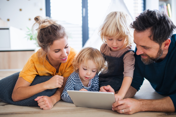 Front view of young family with two small children indoors in bedroom, using tablet.