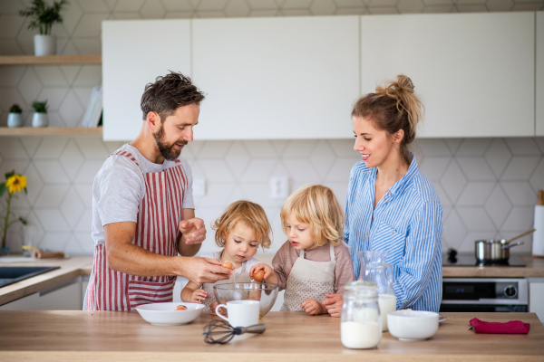 Front view of young family with two small children indoors in kitchen, breaking eggs when cooking.