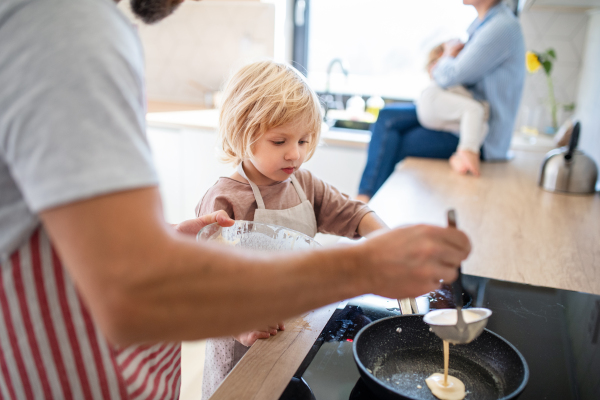 A side view of small boy helping father indoors in kitchen with making pancakes.