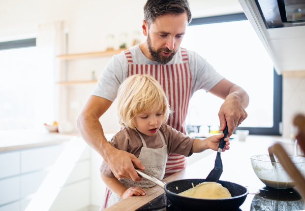 A front view of small boy with father indoors in kitchen making pancakes.