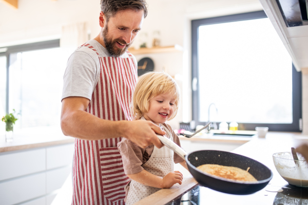 A side view of small boy helping father indoors in kitchen with making pancakes.