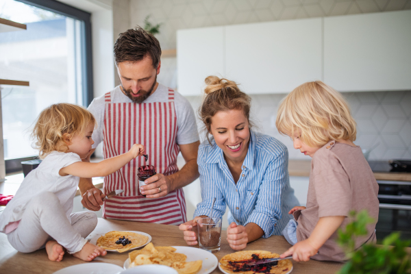 Front view of young family with two small children indoors in kitchen, eating pancakes.