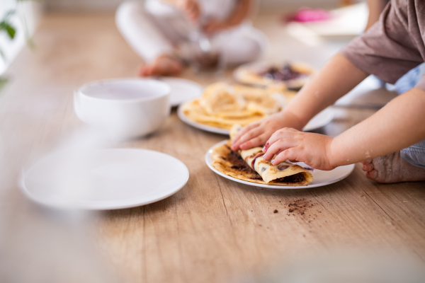 Unrecognizable two small children indoors in kitchen eating pancakes, midsection.