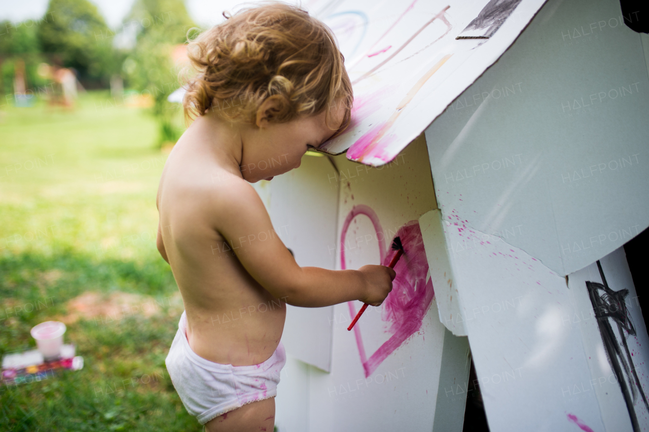 Happy topless small blond girl painting paper house outdoors in summer.