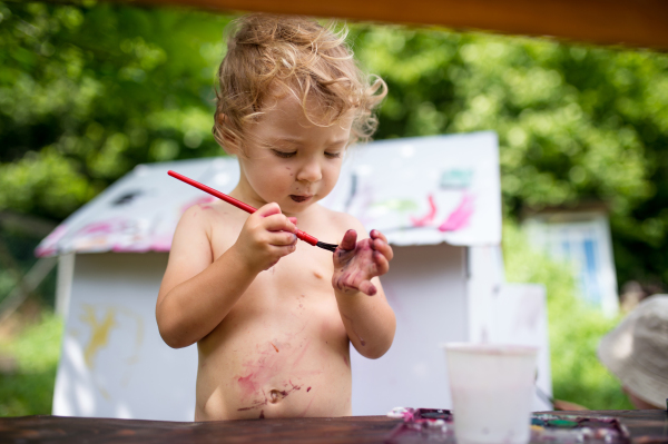 Happy topless small blond girl painting paper house outdoors in summer.