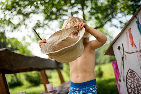 Topless small blond boy with hat painting outdoors in summer, playing.