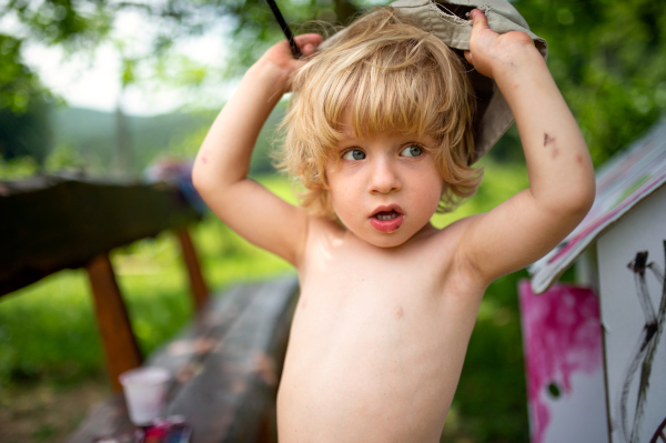 Topless small blond boy with hat painting outdoors in summer, looking at camera.