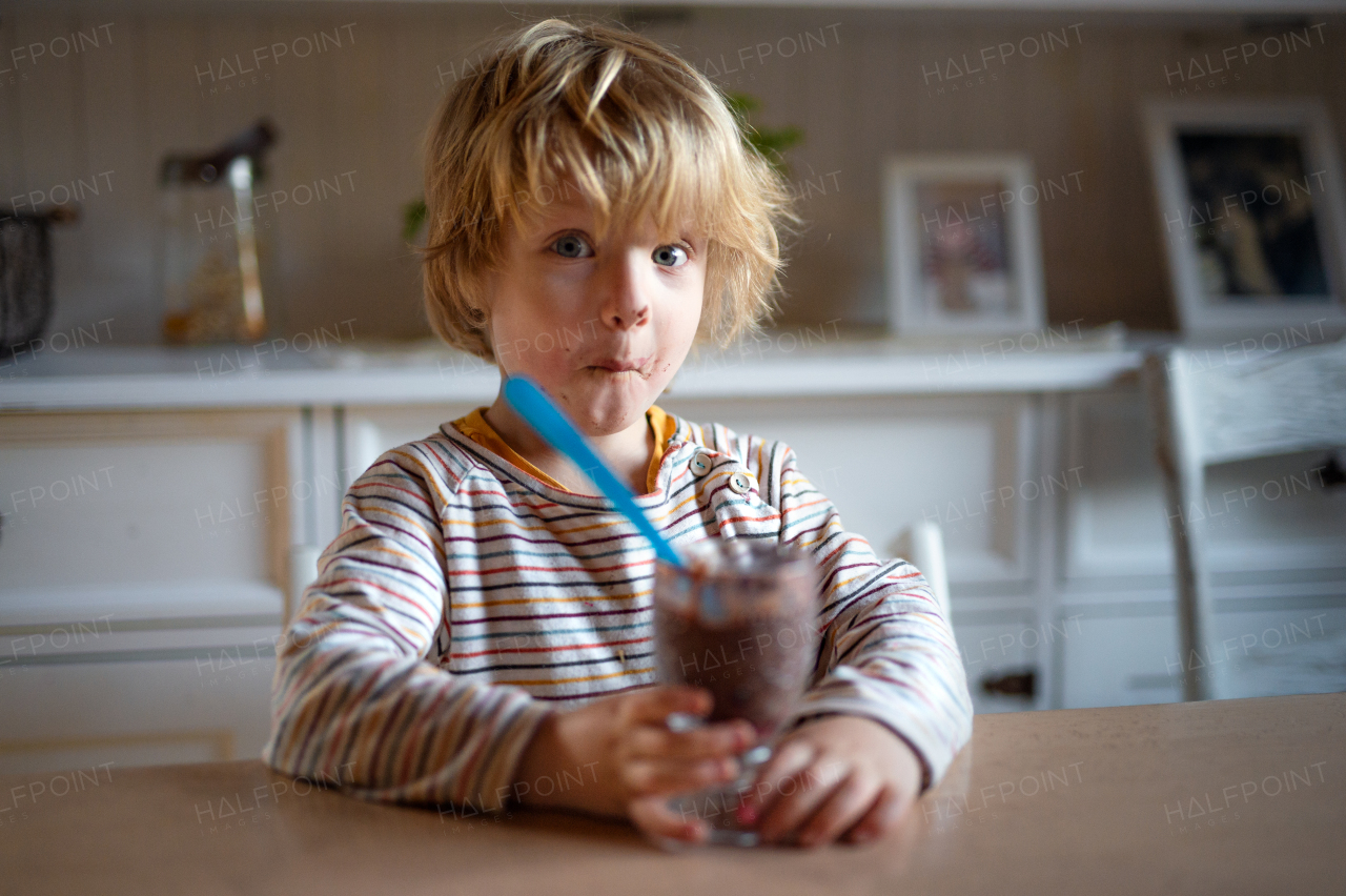 Portrait of small boy with dirty mouth indoors in kitchen at home, eating pudding.