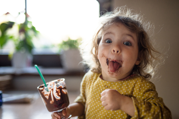 Happy small girl with dirty mouth indoors in kitchen at home, eating pudding.