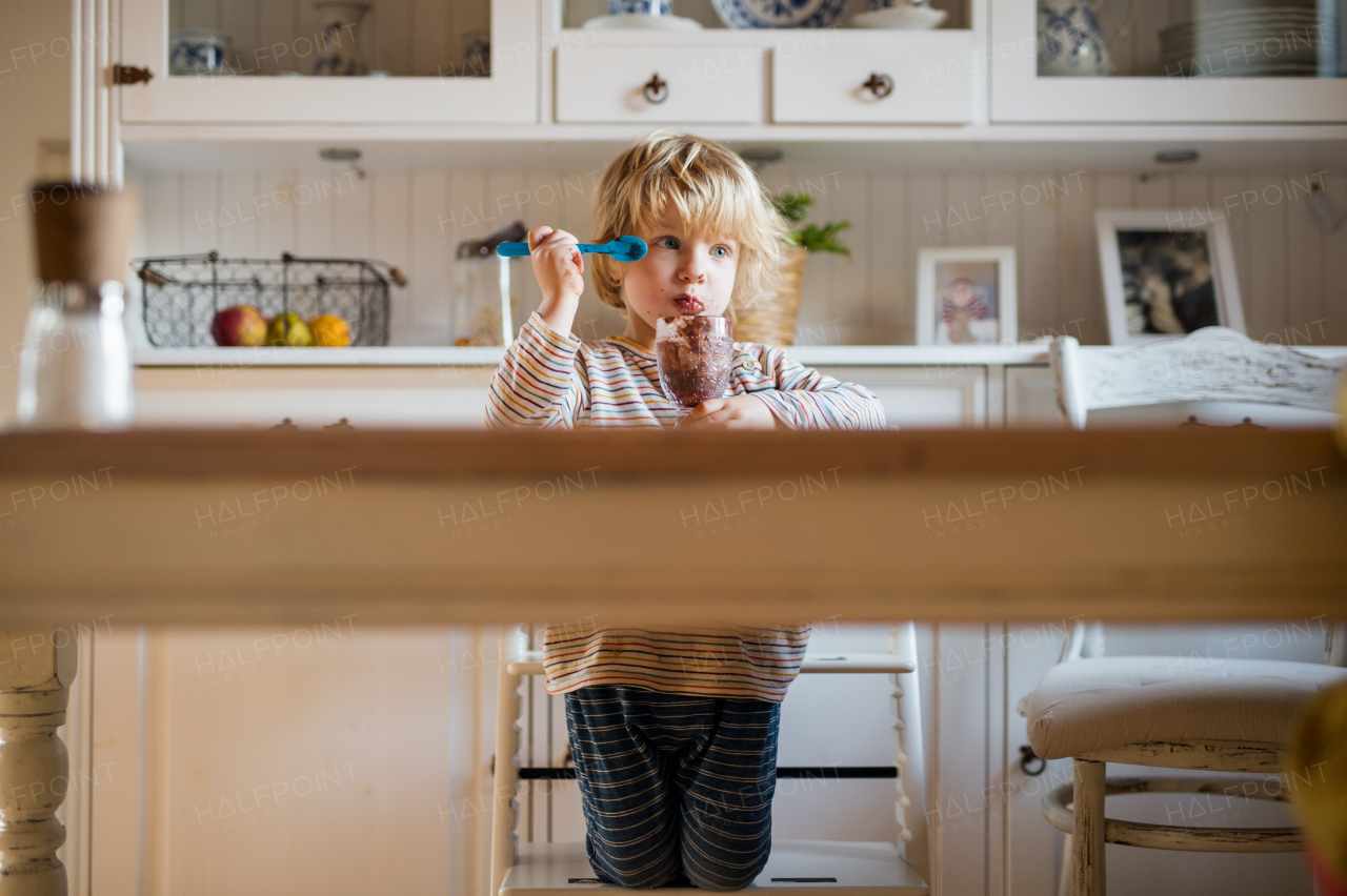 Sad small boy with dirty mouth indoors in kitchen at home, eating pudding.