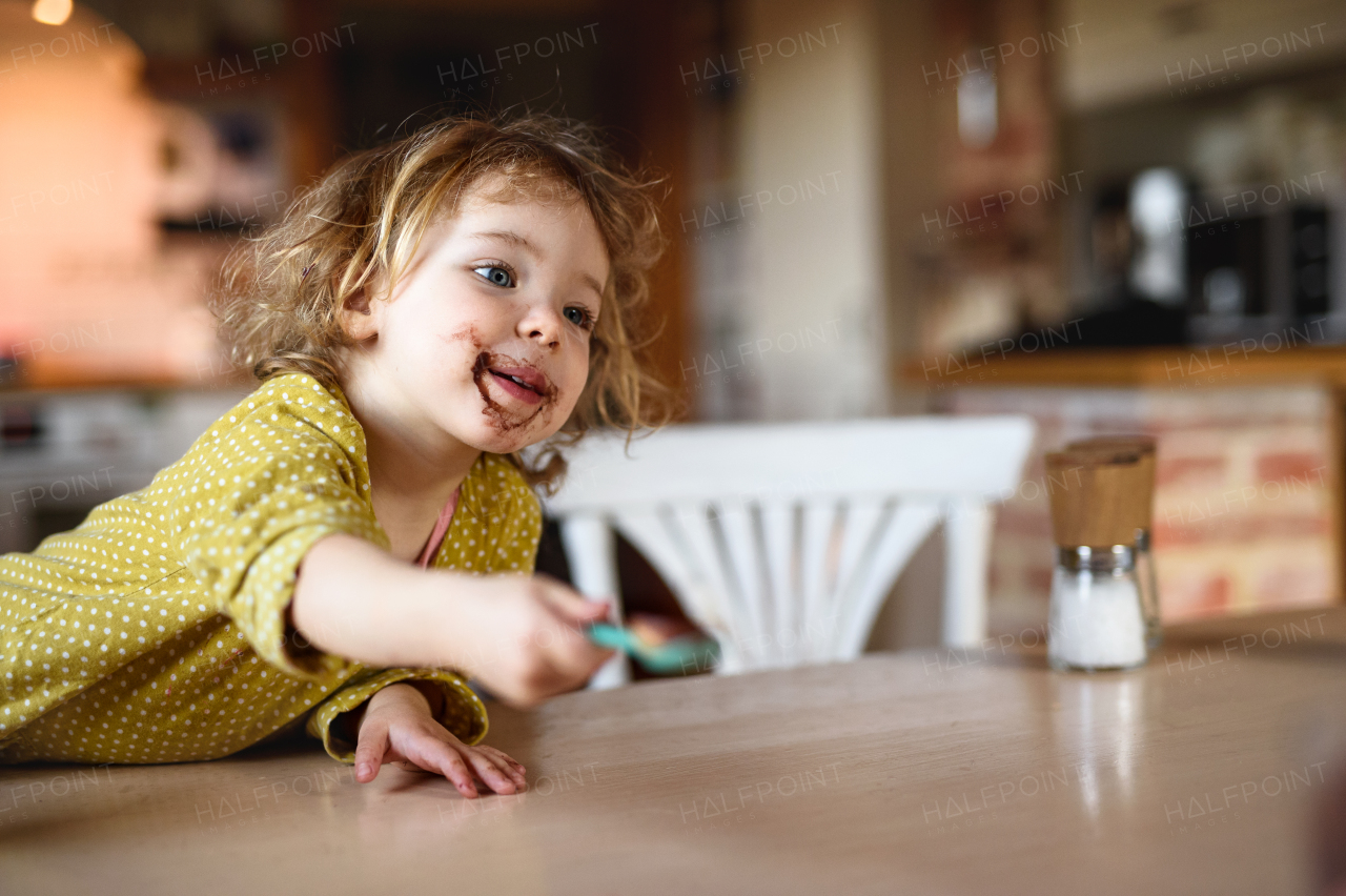 Happy small girl with dirty mouth indoors in kitchen at home, eating pudding.