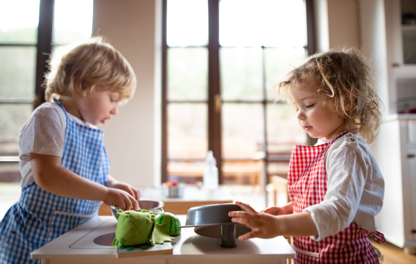 Happy small boy and girl with apron playing indoors with toy kitchen at home.