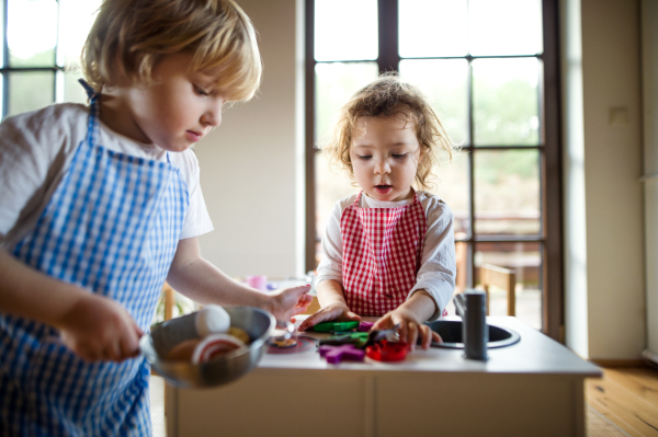 Happy small boy and girl with apron playing indoors with toy kitchen at home.